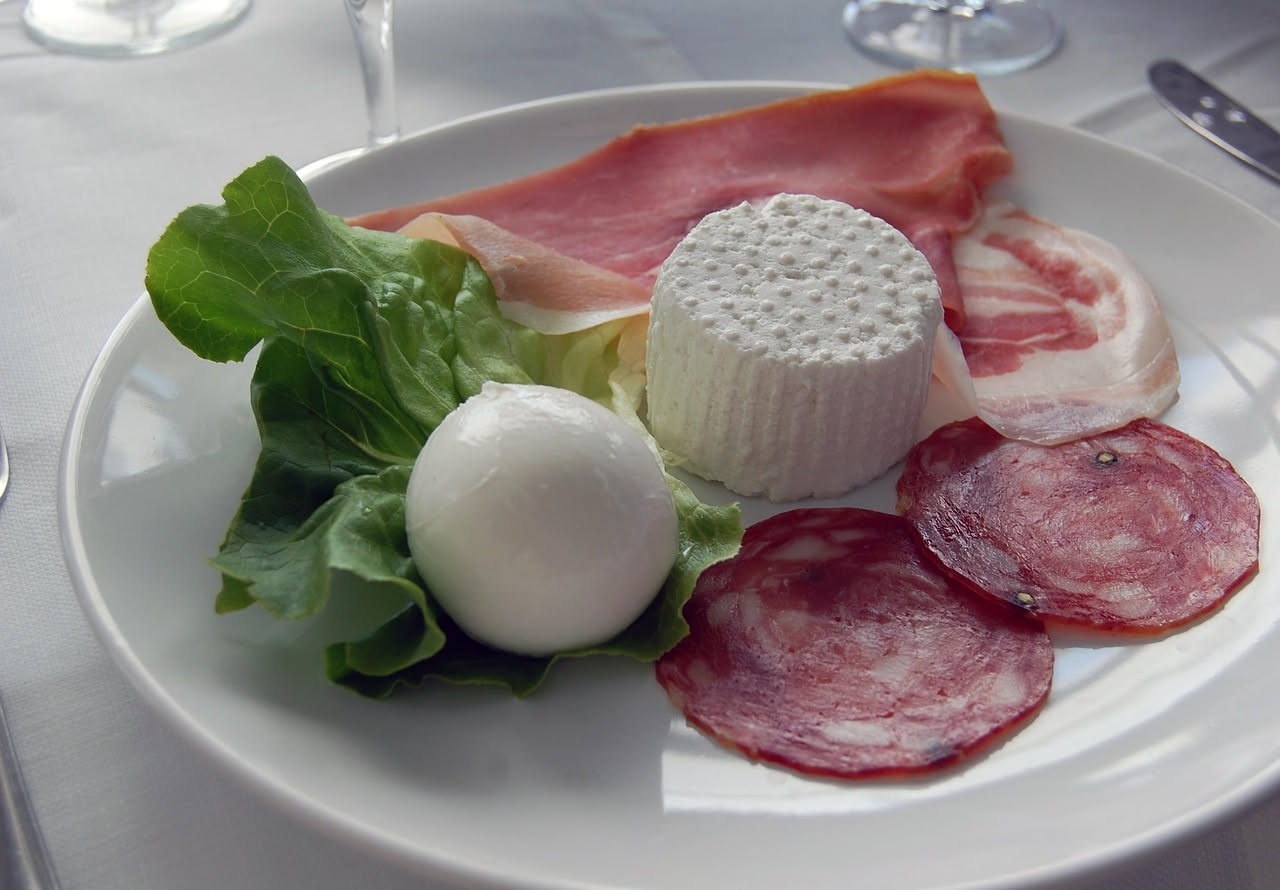 Ciotola di ceramica bianca con jota fredda: fagioli bianchi e rossi, crauti chiari, peperoncino rosso affettato, brodo trasparente color ambra, servita con ghiaccio intorno al piatto